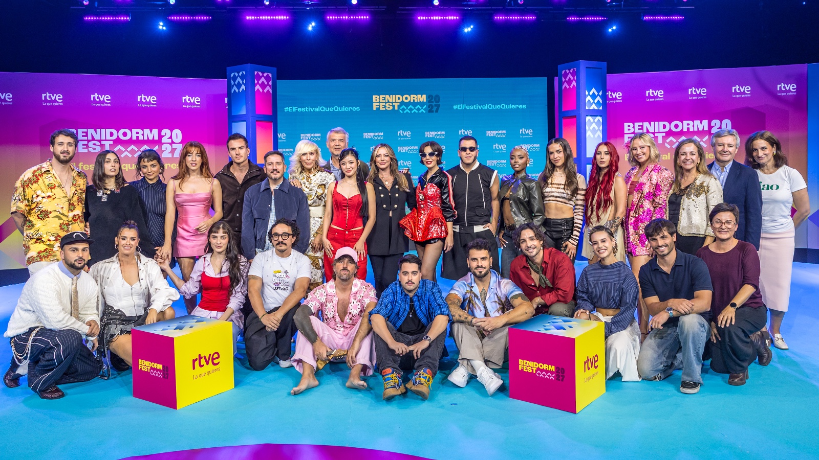Foto de familia de los artistas del Benidorm Fest 2027 posando sonrientes en un plató con una pantalla curva de colores fucsia y violeta.